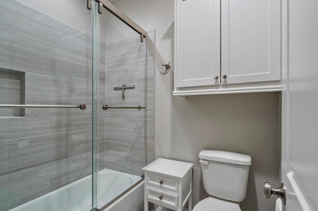 14486 Meadow Glen, Lindale, TX 75771 Bathtub with sliding glass shower door, gray tiled shower surround, recessed shower niche, and wall-mounted white cabinetry