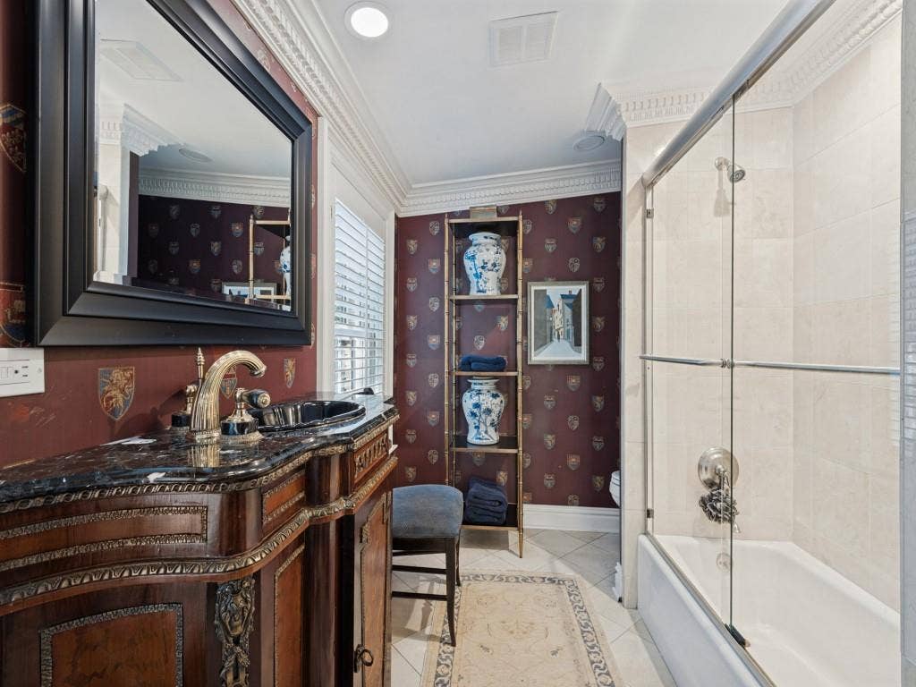 6858 Tokalon Drive, Dallas, TX 75214 Custom vanity with ornate wood detailing, stone countertop, integrated sink, and wall-mounted mirror, featuring crown molding, tile flooring, and a shower-tub combination with a glass enclosure