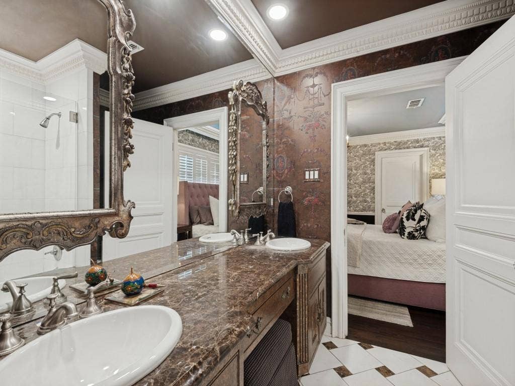 6858 Tokalon Drive, Dallas, TX 75214 Double vanity featuring stone countertops with dual white drop-in sinks, ornate wall-mounted mirrors, custom cabinetry, and patterned wall coverings complemented by detailed crown molding and tile flooring