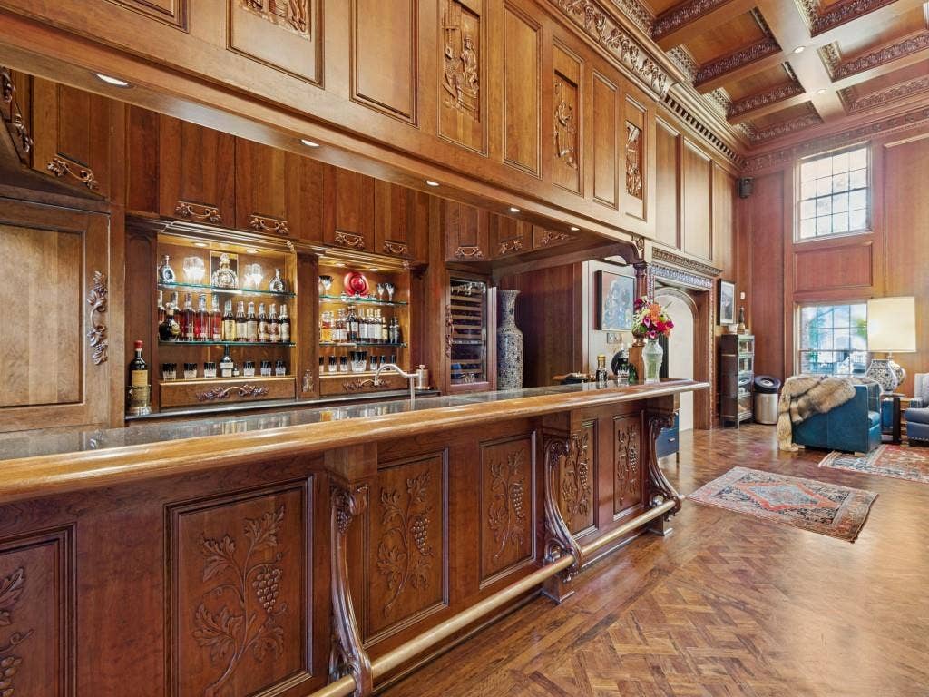 6858 Tokalon Drive, Dallas, TX 75214 Custom wood-paneled bar featuring intricate relief carvings, coffered wood ceilings, built-in glass shelving, a metal foot rail, and herringbone-pattern wood flooring