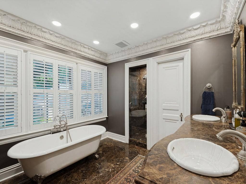6858 Tokalon Drive, Dallas, TX 75214 Freestanding clawfoot tub with bridge-style faucet, dual-sink vanity with stone countertop, walk-in stone-tiled shower, crown molding, and plantation shutters