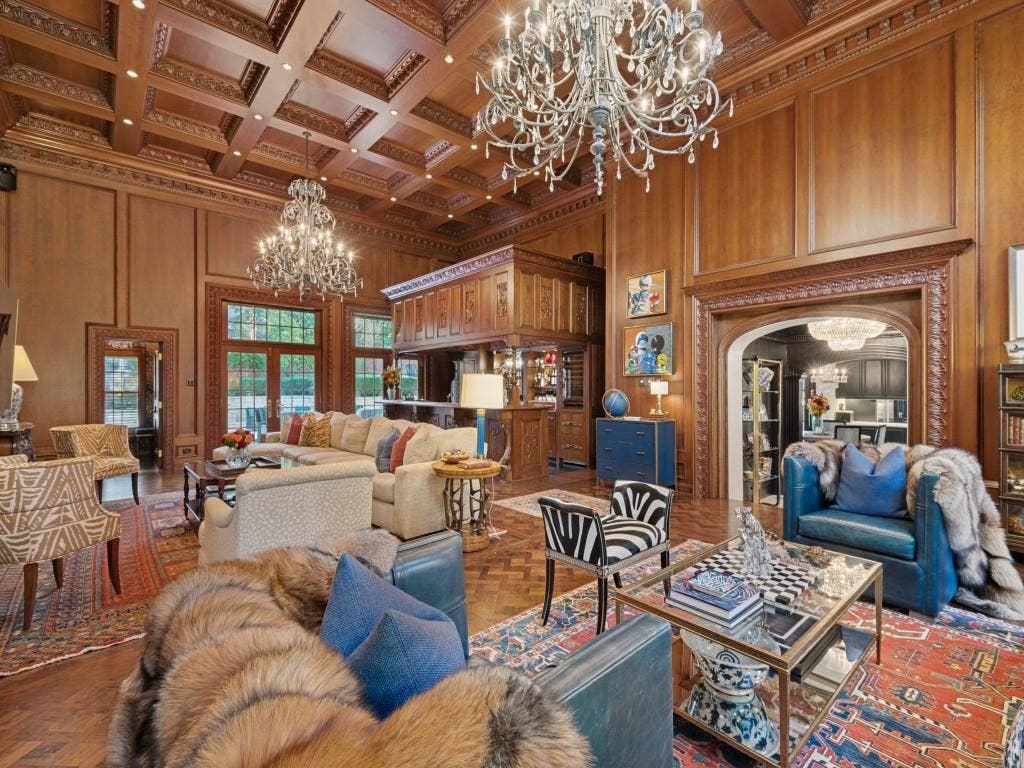 6858 Tokalon Drive, Dallas, TX 75214 Grand room featuring extensive wood-paneled walls, a coffered ceiling with intricate detailing, polished herringbone wood-finish flooring, two large crystal chandeliers, and a custom built-in bar