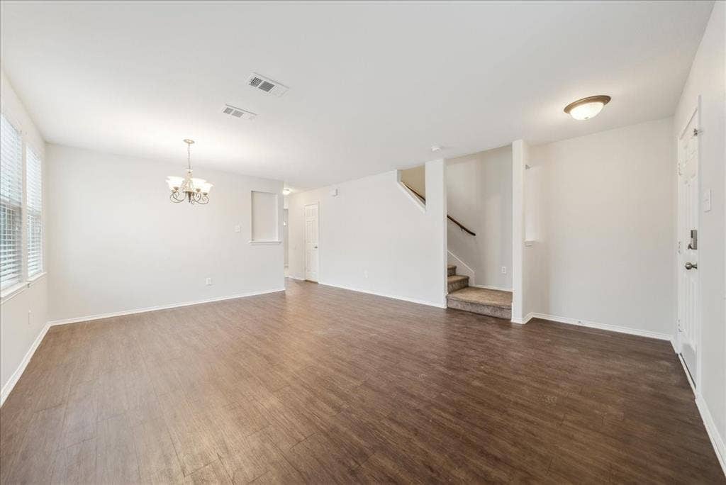 Wood-finish flooring throughout, bright white walls, multiple windows with horizontal blinds, overhead chandelier, and a carpeted staircase