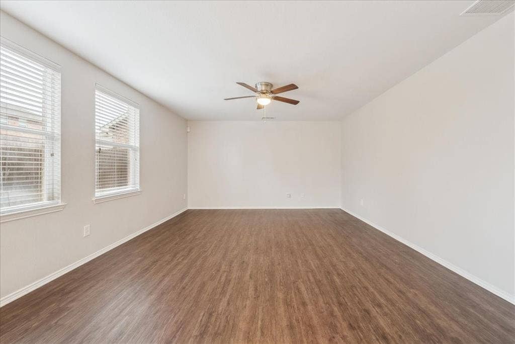 Spacious room featuring wood-finish flooring, multiple windows with blinds, a ceiling fan, and neutral paint