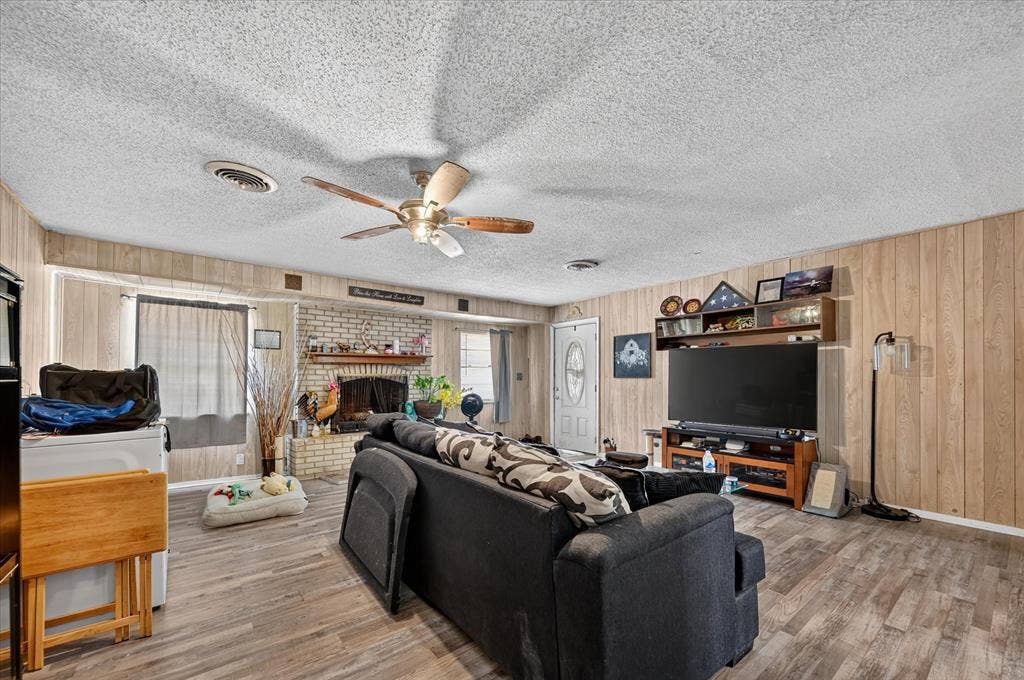 3320 Shorewood Drive, Mesquite, TX 75150 Living area featuring wood-finish flooring, paneled walls, a ceiling fan, and an exposed brick fireplace