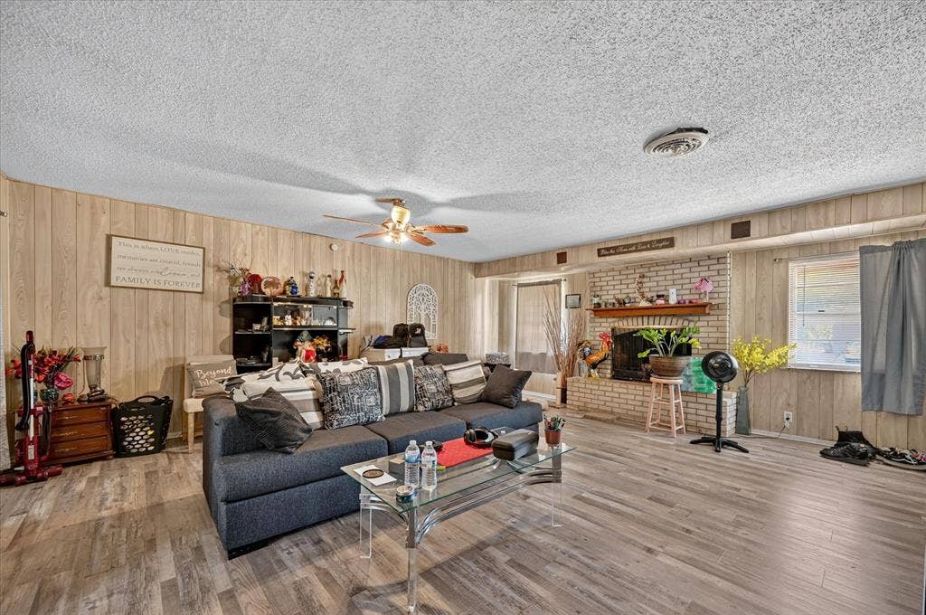 3320 Shorewood Drive, Mesquite, TX 75150 Expansive living area featuring wood-finish flooring, paneled wall accents, a brick-surround fireplace with a mantel, and an overhead ceiling fan