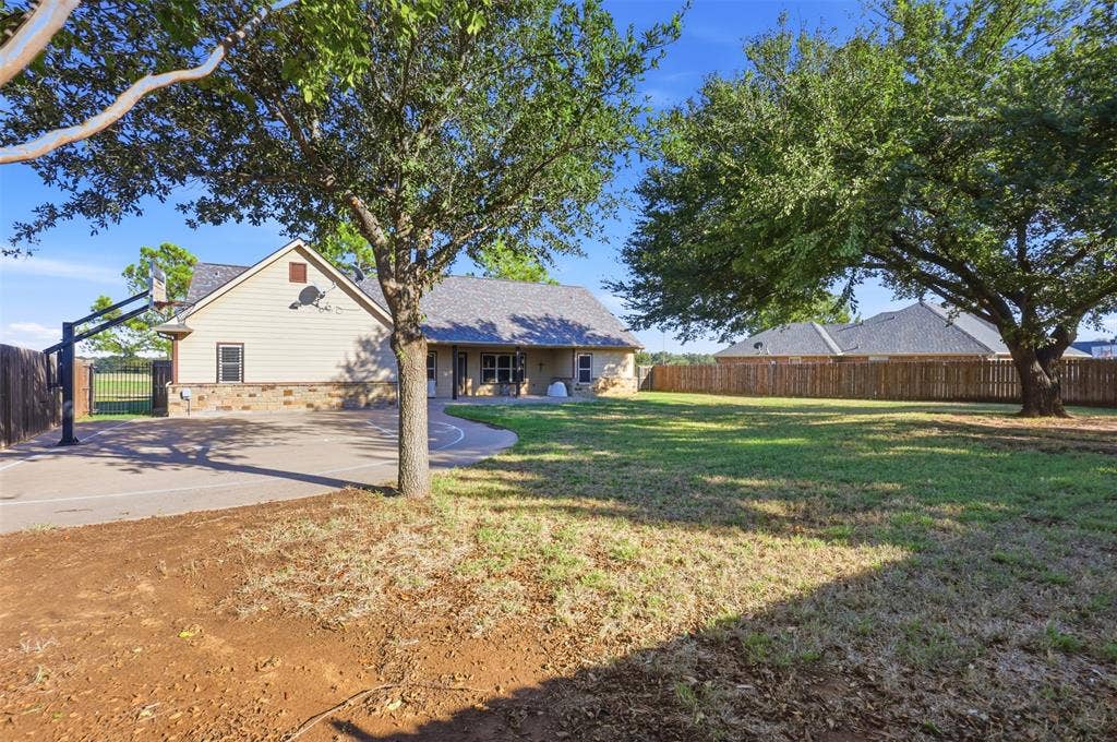 Expansive backyard with a manicured lawn, mature trees, wood fence, and a paved sport court
