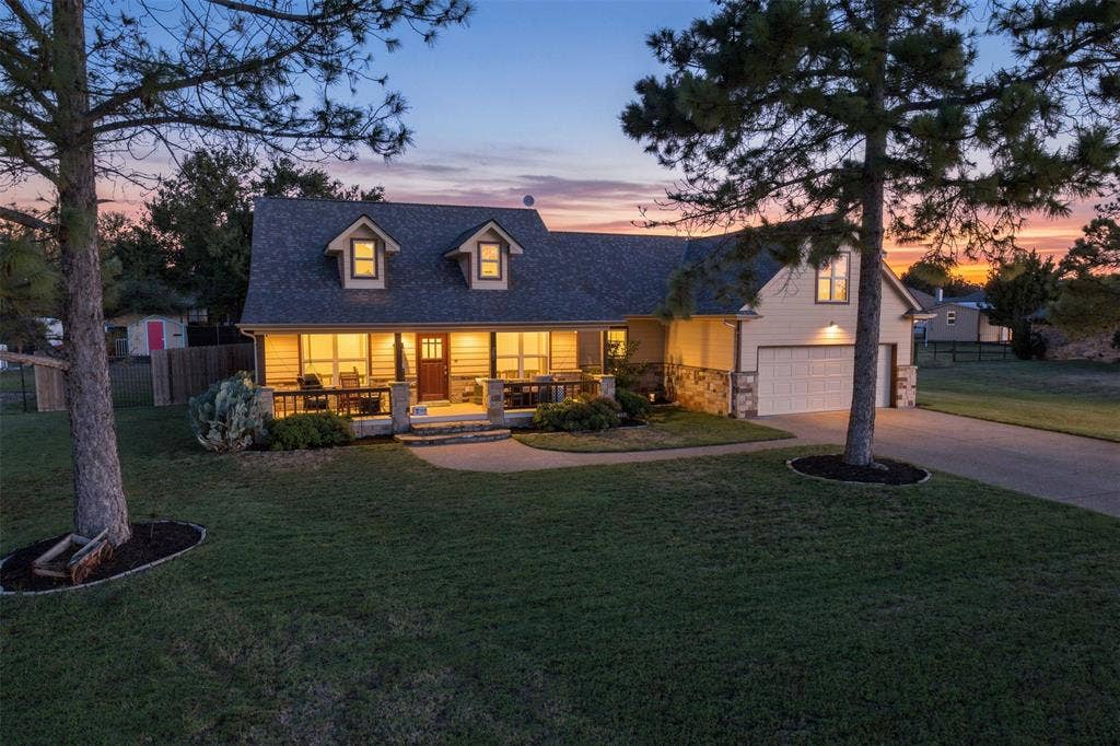 Gable roofline with dormer windows, stone accent siding, covered front porch, attached garage, and paved driveway