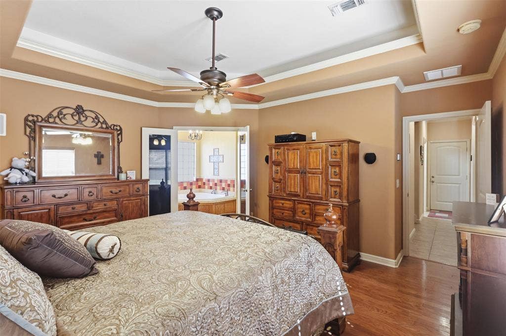 Recessed tray ceiling with crown molding, wood-finish flooring, ceiling fan with integrated lighting, and an alcove with a built-in jetted tub
