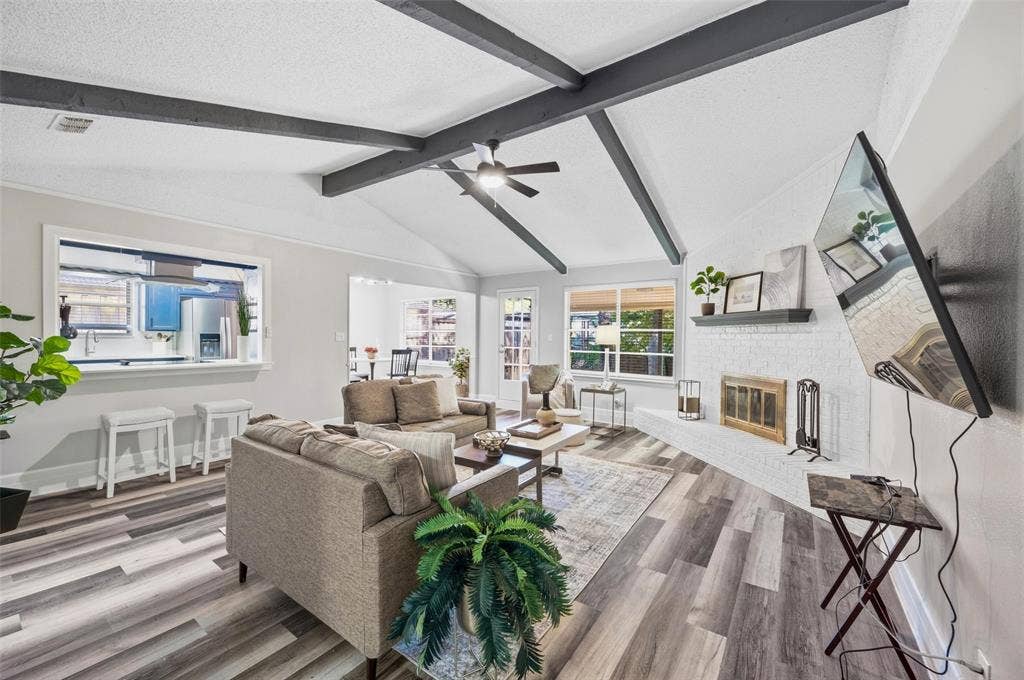 Vaulted ceiling with exposed beams, wood-finish flooring, white brick fireplace with a mantel, and a kitchen pass-through window
