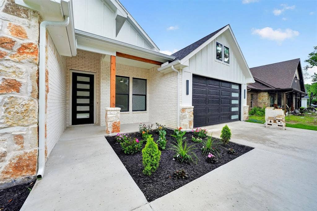 120 Springwood Road, Enchanted Oaks, TX 75156 White brick exterior with stone accents, a modern black entry door, a dark garage door with horizontal windows, and a landscaped front planter