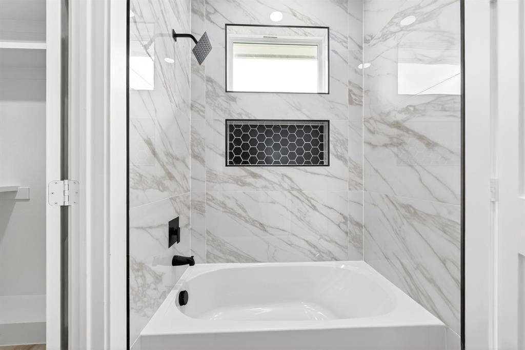 120 Springwood Road, Enchanted Oaks, TX 75156 Bathtub with white porcelain finish, matte black fixtures, marble-look tiled surround, recessed shower niche with black hexagonal tile, and integrated window