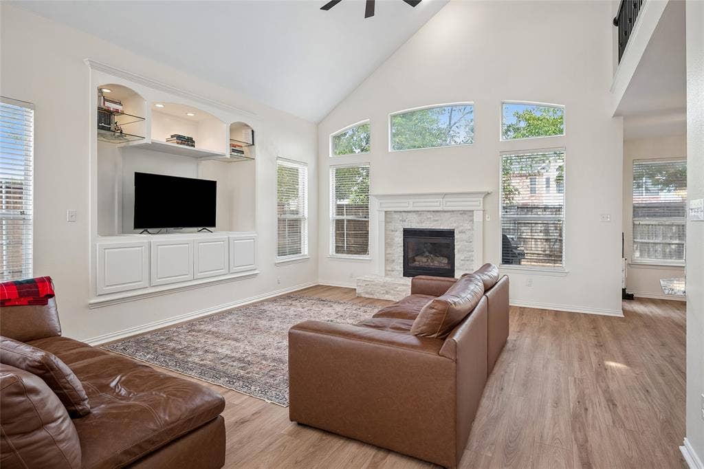 3801 Kite Meadow Drive, Plano, TX 75074 Vaulted ceiling with a ceiling fan, built-in entertainment center with shelving, fireplace with a tiled surround and mantel, abundant natural light from multiple windows, and wood-finish flooring
