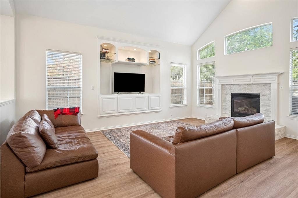 3801 Kite Meadow Drive, Plano, TX 75074 Vaulted ceiling with high windows, built-in media center with shelving, white stone-surround fireplace with a classic mantel, and wood-finish flooring