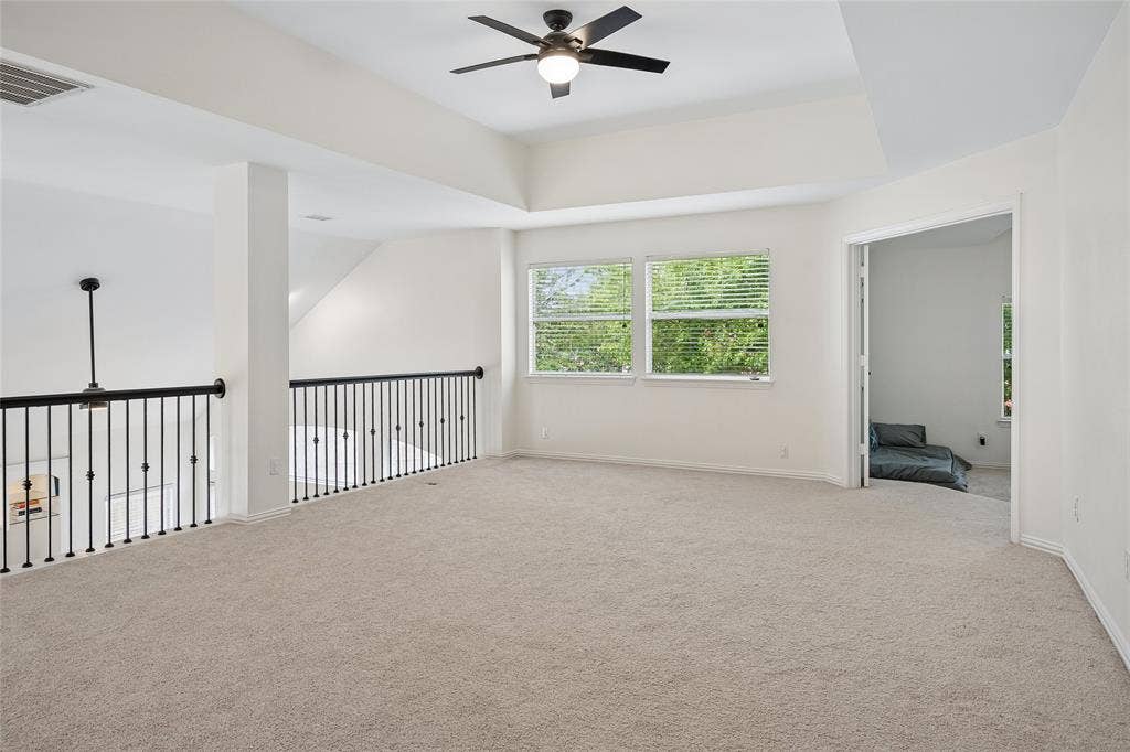 3801 Kite Meadow Drive, Plano, TX 75074 Spacious carpeted loft featuring a tray ceiling with a ceiling fan, multiple windows with blinds, and wrought iron railings overlooking the floor below