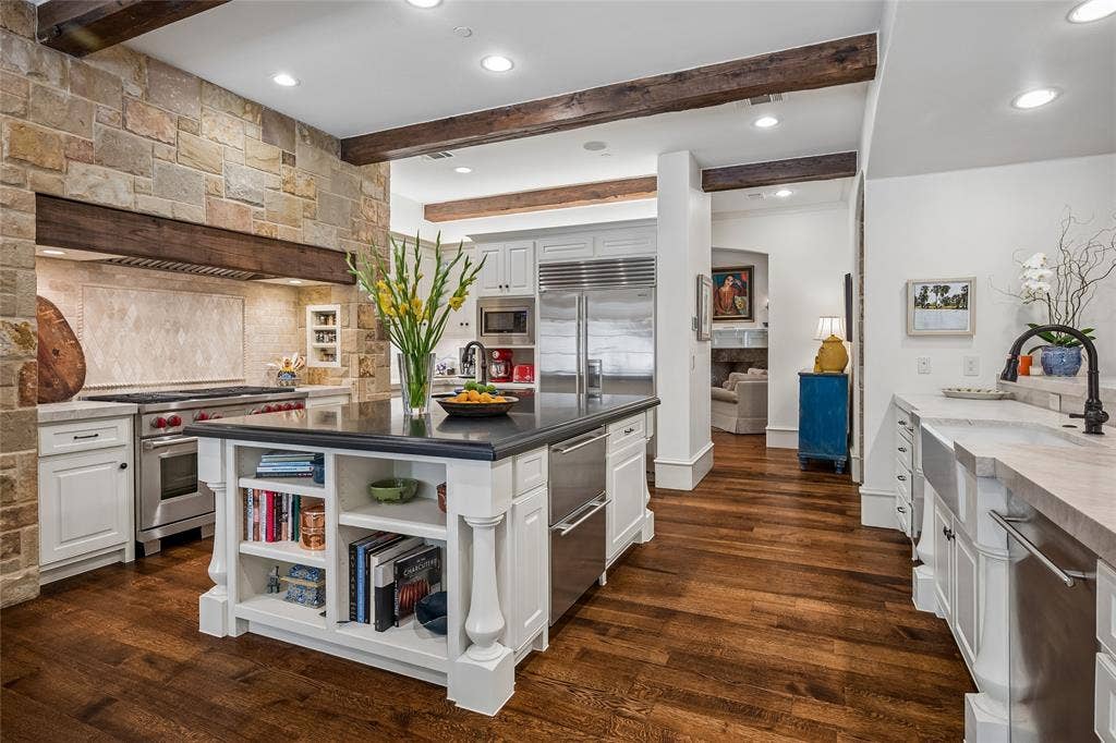 5222 Stonegate Road, Dallas, TX 75209 Gourmet kitchen featuring a stone accent wall, wood beams, stainless steel appliances, a center island with built-in shelving, and wood-finish flooring