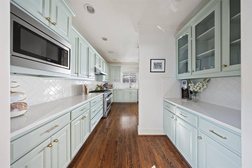 5709 Kenwood Avenue, Dallas, TX 75206 Recessed lighting illuminates the galley-style kitchen featuring light-toned cabinetry, white herringbone tile backsplash, integrated stainless steel microwave, and wood-finish flooring