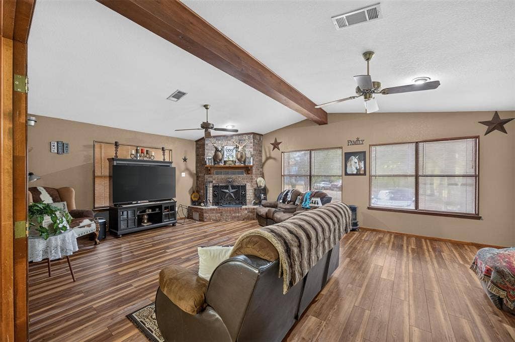 Expansive living area featuring wood-finish flooring, a brick fireplace, and exposed ceiling beams