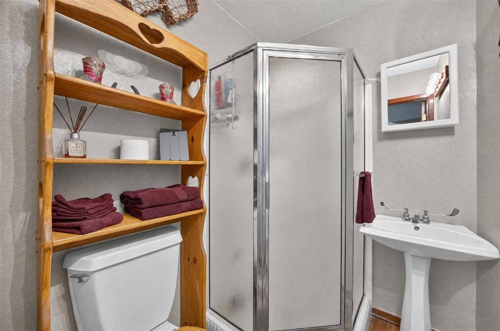 Corner shower with frosted glass enclosure, pedestal sink, wall-mounted mirror, and wood shelving unit
