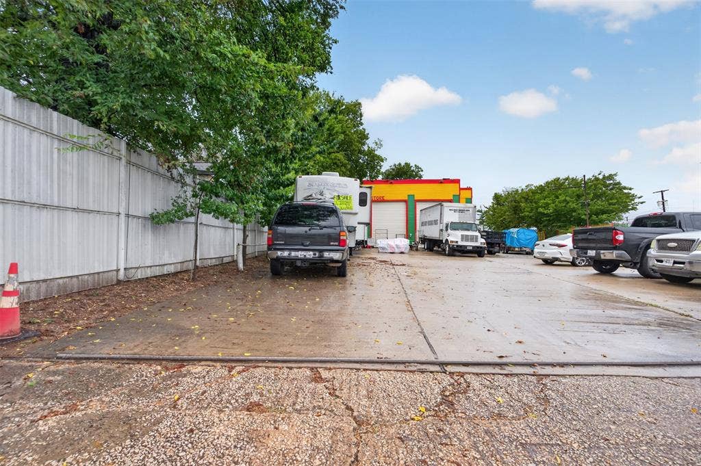 2301 S Denley Drive, Dallas, TX 75216 Paved parking area featuring a corrugated metal fence, mature trees, and a concrete surface