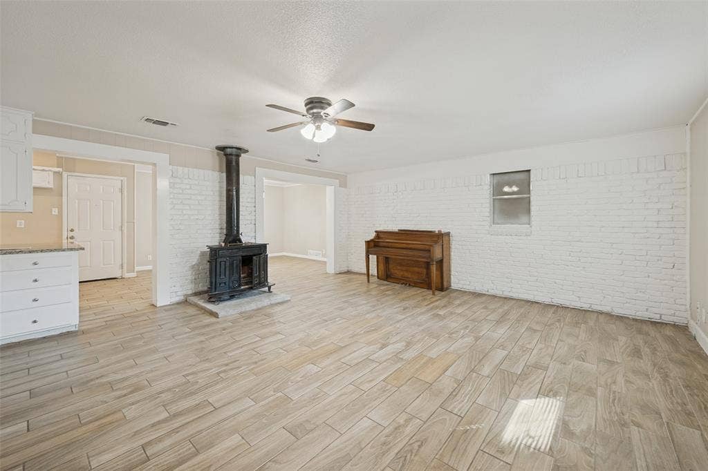 213 W Dorris Drive, Grand Prairie, TX 75051 Wood-finish flooring, white brick accent wall, wood-burning stove, ceiling fan, and white cabinetry with granite-style countertops