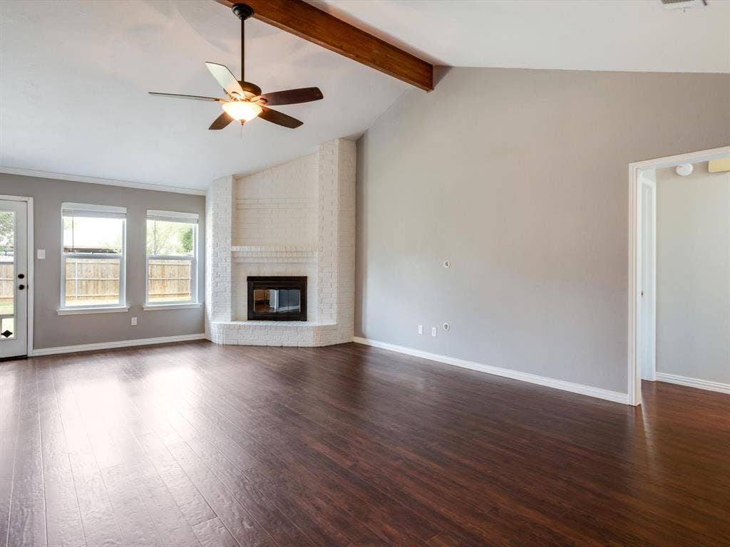 Vaulted ceiling featuring an exposed wood beam, corner white-painted brick fireplace, wood-finish flooring, and a ceiling fan with integrated light fixture