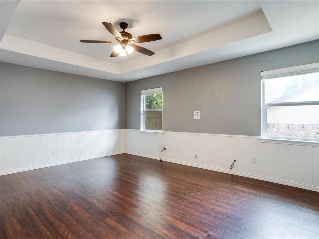 Tray ceiling featuring a central multi-blade ceiling fan with integrated lighting, wood-finish flooring, and white wainscoting