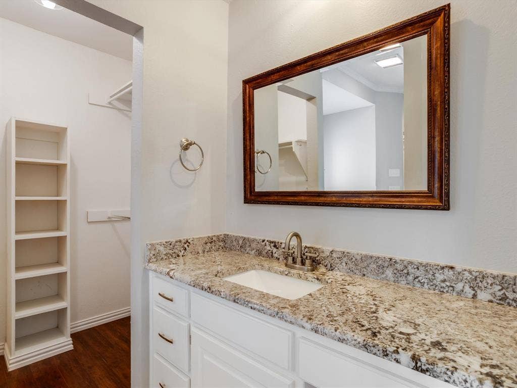 Vanity featuring a stone-look countertop, white shaker-style cabinetry, an integrated rectangular sink with a brushed metal faucet, a framed mirror, and built-in shelving