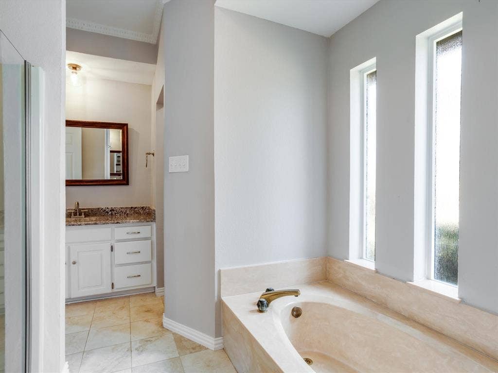 Soaking tub featuring a stone-tile surround, dual vertical windows, light-tone tile flooring, and a built-in vanity with stone-surfaced countertop