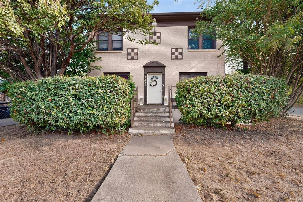 Brick exterior featuring decorative square accents, mature trees, and established perimeter hedging