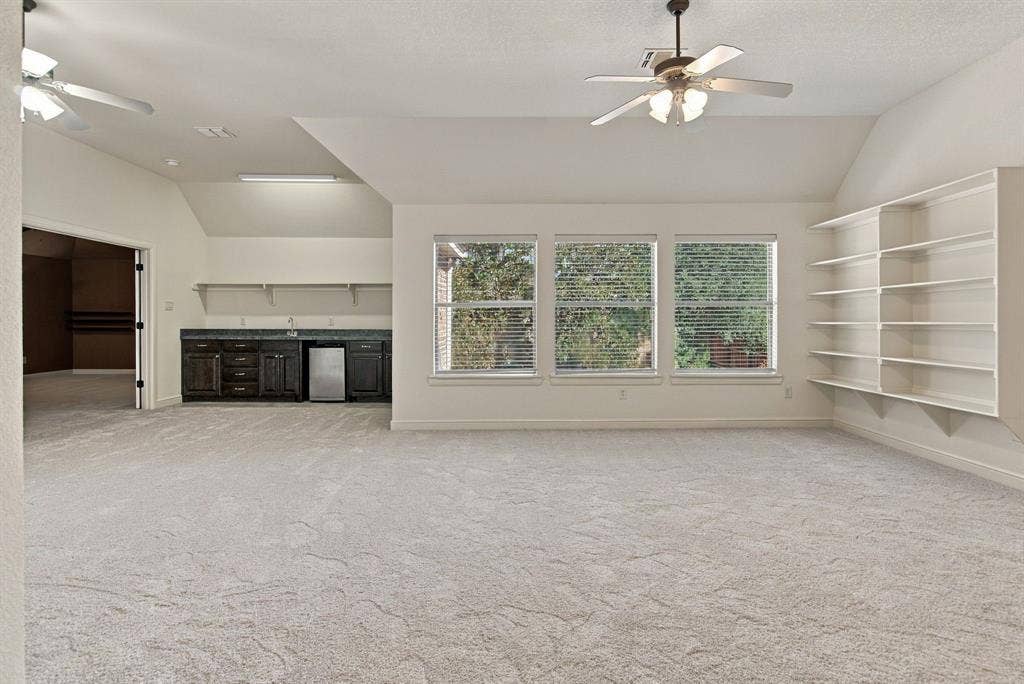 7004 Cherrybark Lane, North Richland Hills, TX 76182 Expansive room featuring light-colored carpeting, multiple windows, built-in shelving, and a wet bar with dark cabinetry and a compact refrigerator