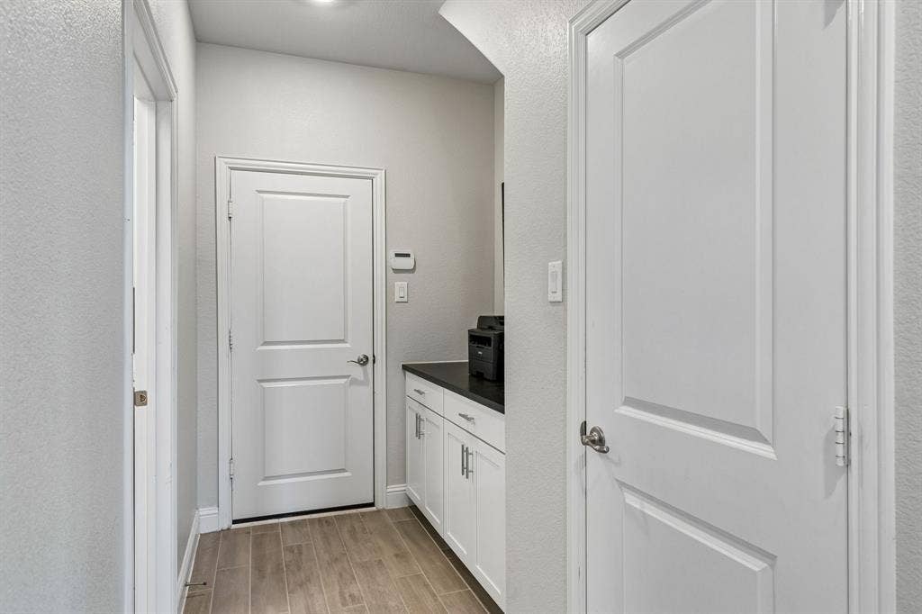 3707 Roth Drive, McKinney, TX 75071 Mud Room showcases crisp white paneled doors, soft neutral walls, warm wood-look flooring, and beautifully appointed built-in cabinetry with a sleek dark countertop, creating a polished and highly functional transition into the home's utility spaces.