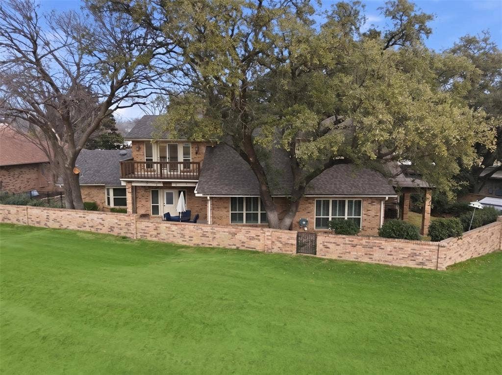 4424 Westridge Avenue, Fort Worth, TX 76116 Brick exterior with a second-story balcony, expansive green lawn, mature trees, and a brick privacy wall with an ornamental gate