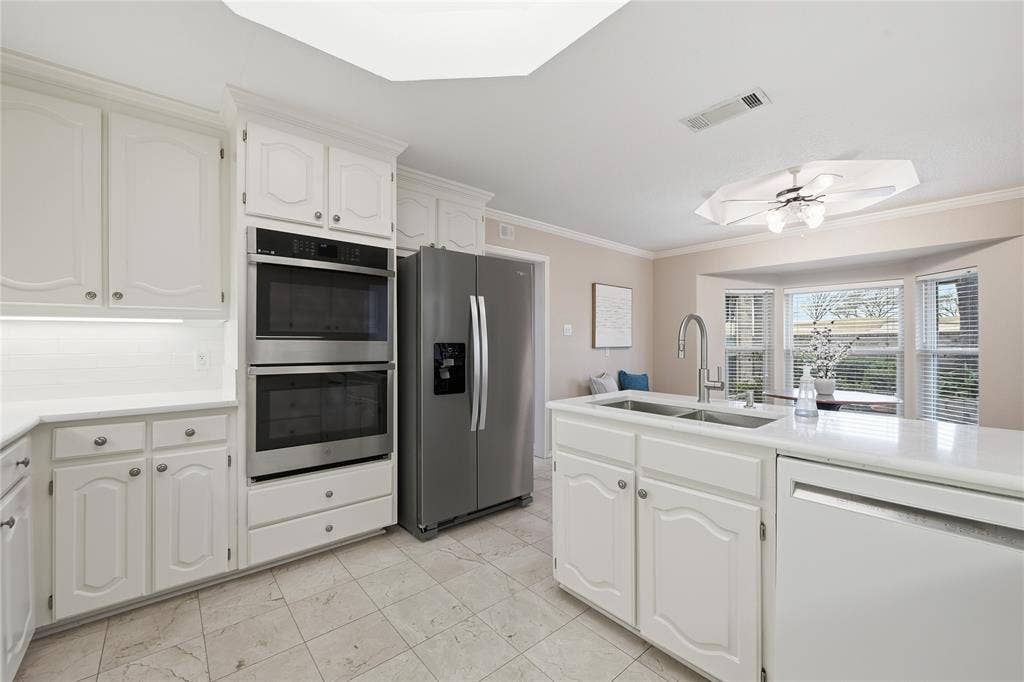 4424 Westridge Avenue, Fort Worth, TX 76116 White cabinetry with crown molding, stainless steel double wall ovens, a bay window, a dual basin undermount sink with a gooseneck faucet, and light tile flooring