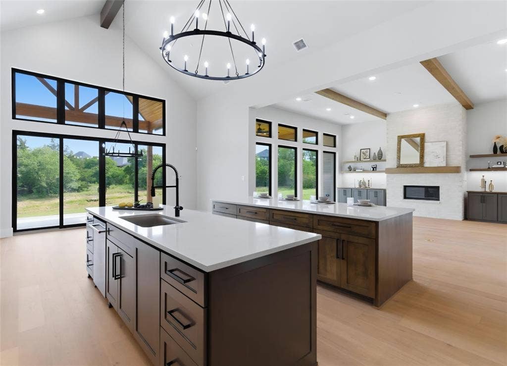 2608 Southbay, Northlake, TX 76226 Vaulted ceilings with exposed beams, multiple kitchen islands with white countertops and dark cabinetry, wood-finish flooring, and a fireplace with a white brick surround and wood mantel