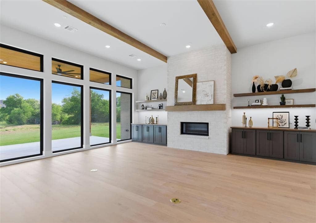 2608 Southbay, Northlake, TX 76226 Open-concept living space with full-height windows, wood-finish flooring, a white brick fireplace, exposed ceiling beams, and built-in cabinetry with floating shelves
