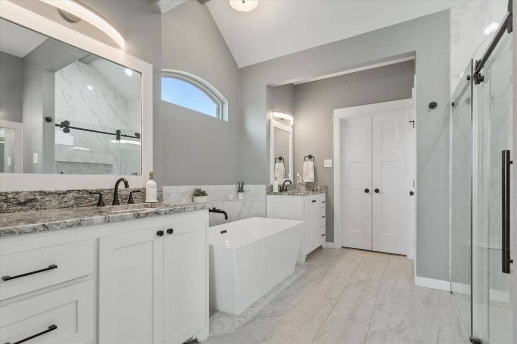 10305 Creekside Lane, Waco, TX 76712 Dual vanities with stone countertops, a freestanding soaking tub, a walk-in shower with a sliding glass door, an arched transom window, and vaulted ceilings