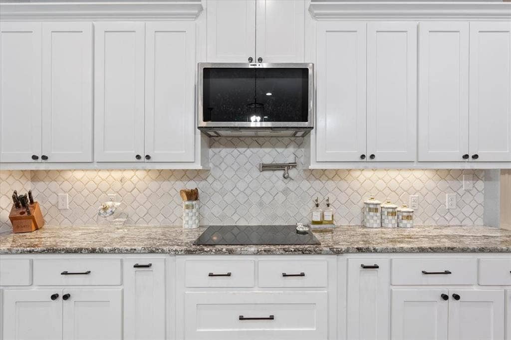 10305 Creekside Lane, Waco, TX 76712 White shaker-style cabinetry, patterned tile backsplash, built-in stainless steel microwave, stone countertops, and black electric cooktop