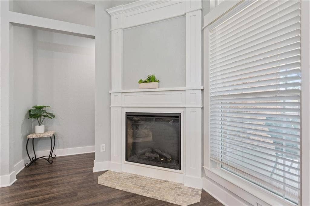 10305 Creekside Lane, Waco, TX 76712 Corner fireplace with a white mantel and tile hearth, accompanied by wood-finish flooring and a window with white blinds