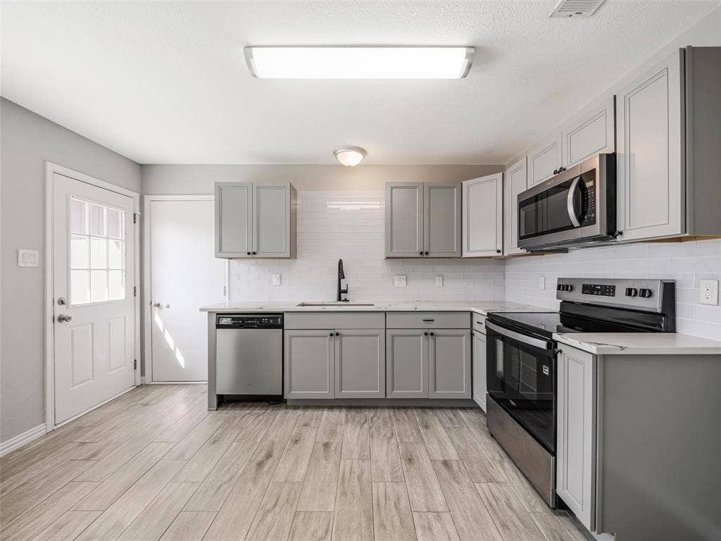 Kitchen featuring light wood-finish flooring, grey cabinetry, white subway tile backsplash, stainless steel appliances, and a glass-paned exterior door