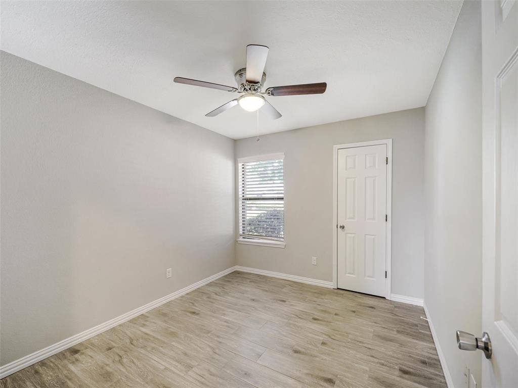 Neutral wall color, wood-finish flooring, ceiling fan with integrated lighting, window with horizontal blinds, and a paneled door