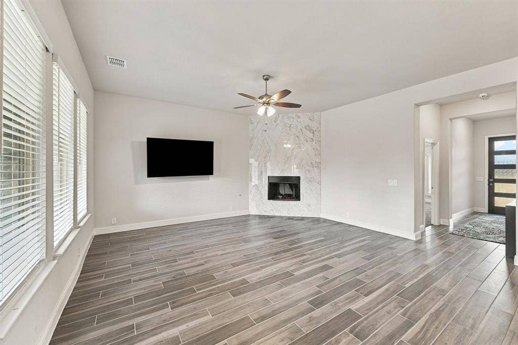 Spacious living area featuring wood-finish tile flooring, a corner fireplace with a marble-finish surround, multiple windows with blinds, and a ceiling fan