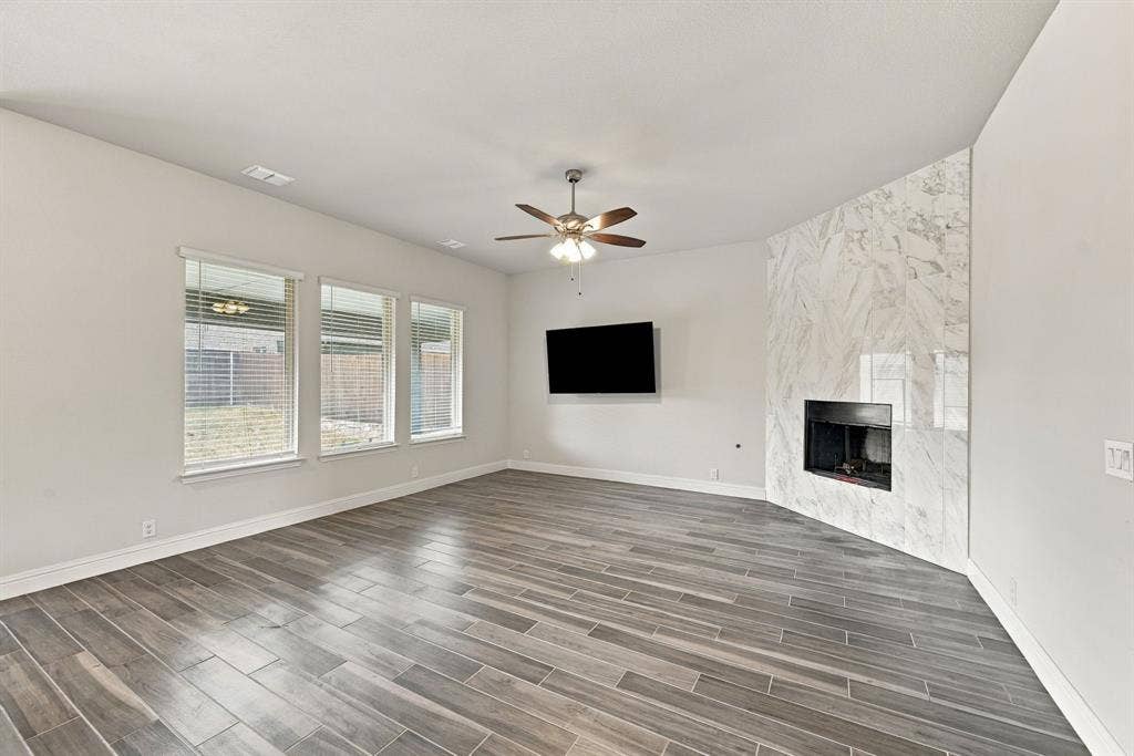 Spacious living area featuring wood-look tile flooring, three large windows with blinds, a ceiling fan, and a fireplace with a light-toned tile surround