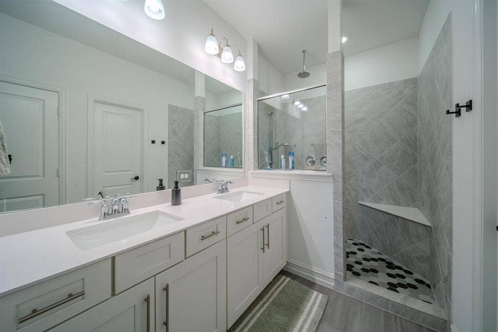 4629 Golden Gate Way, Garland, TX 75043 Dual vanity with undermount sinks and chrome fixtures, large frameless mirror, and walk-in shower with tiled walls, built-in bench, and rain showerhead