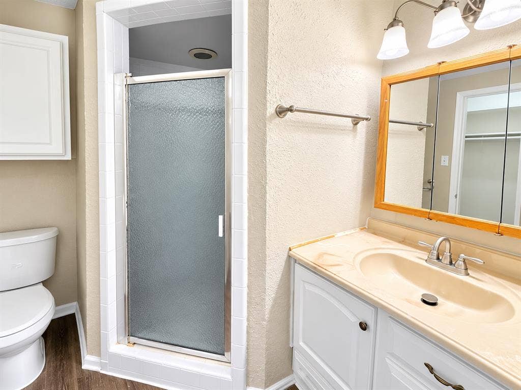 1500 Sherrye Drive, Plano, TX 75074 Wood-finish flooring with white baseboards, shower enclosure with frosted glass door, white toilet, vanity with integrated sink and countertop, and a medicine cabinet with a wood-finish frame