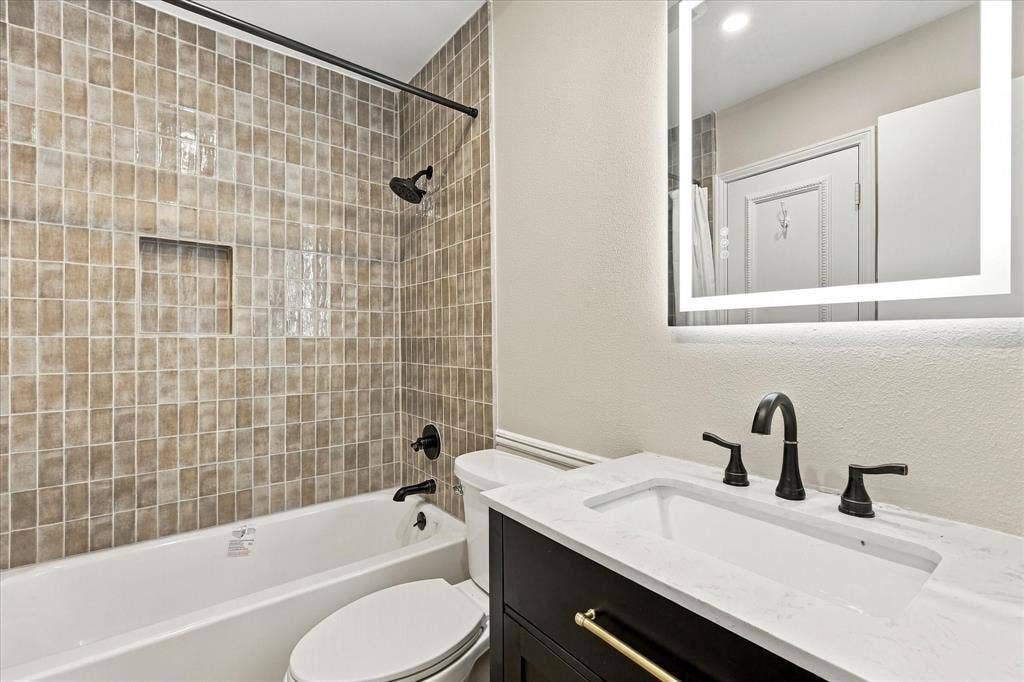 2811 Reedcroft Drive, Farmers Branch, TX 75234 Bathroom featuring a tiled shower surround with a built-in niche, a white tub, a dark wood-finish vanity with gold-tone hardware, a white countertop, and an illuminated mirror