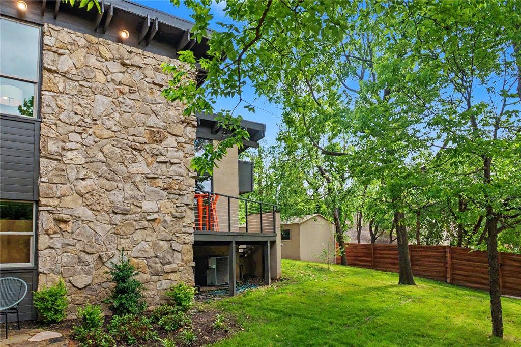 11737 Rogue Way, Dallas, TX 75218 Multi-story exterior featuring natural stone masonry, elevated deck with horizontal rail system, large-pane windows, and landscaped grounds with mature trees and American cedar fencing