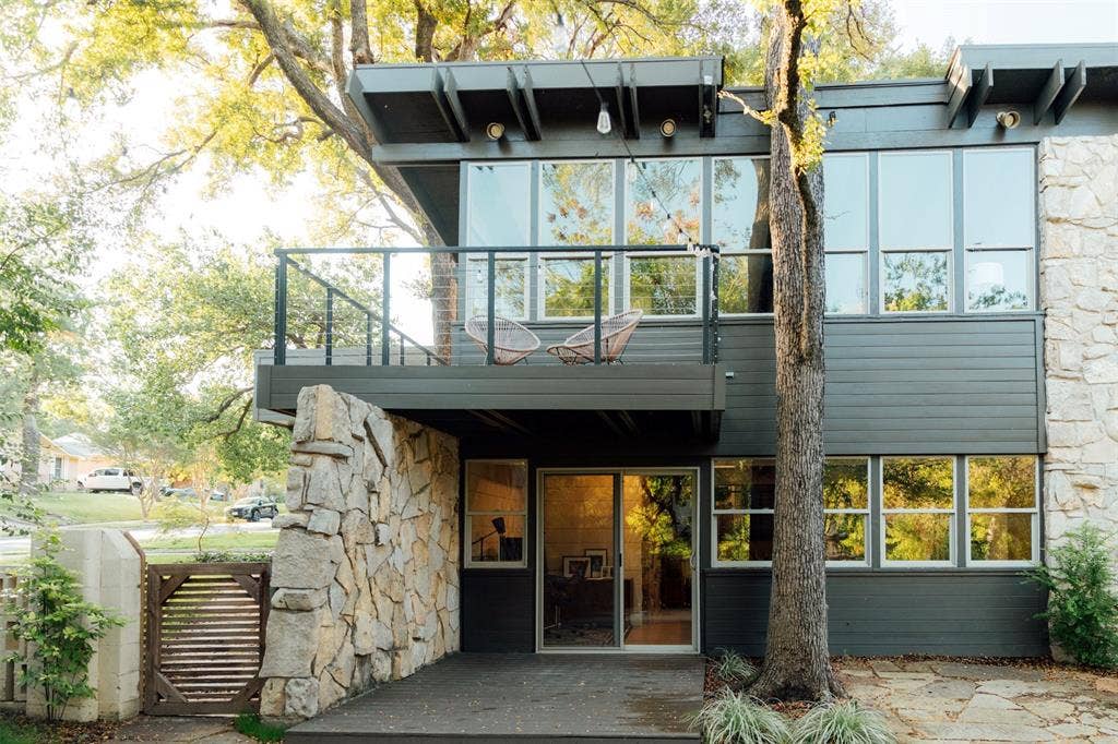 11737 Rogue Way, Dallas, TX 75218 Mid-century modern exterior featuring natural stone masonry, treehouse balcony, expansive floor-to-ceiling windows, and a covered lower-level walkout patio.