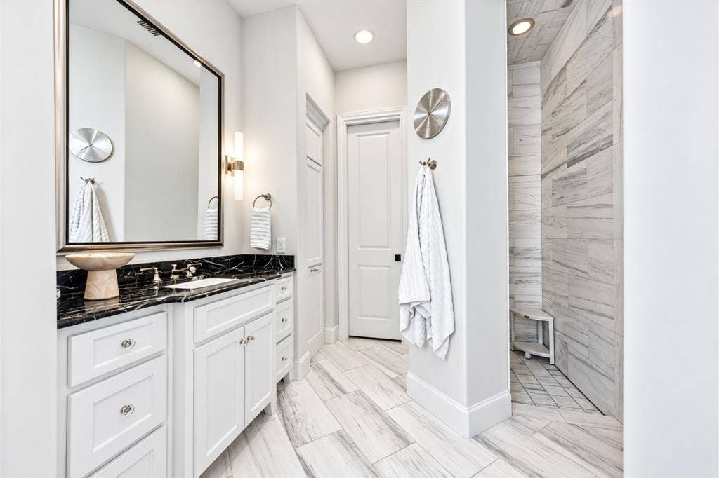 6816 Cousteau Court, Plano, TX 75024 White vanity with a dark countertop and integrated sink, framed mirror with sconce lighting, and a tiled shower with a built-in bench