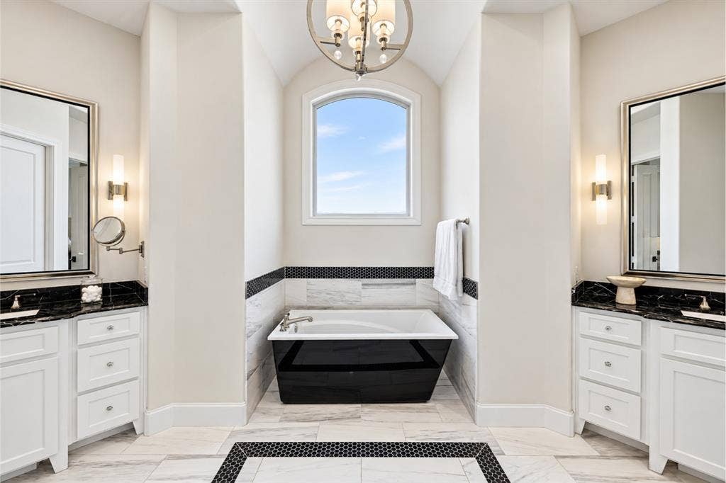 6816 Cousteau Court, Plano, TX 75024 Dual vanity sinks with dark countertops, a standalone soaking tub with dark exterior, arched window, and large format tiled flooring with a mosaic inlay